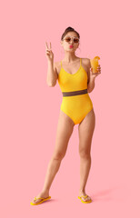 Pretty young woman in swimsuit holding glass of orange juice with straw and showing victory gesture...