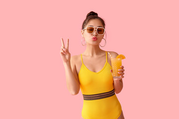 Pretty young woman in swimsuit holding glass of orange juice with straw and showing victory gesture...