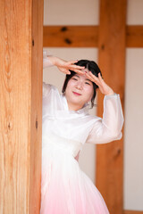 A Korean woman in her twenties wearing a charming pink hanbok peeks from the shadow of a wooden...