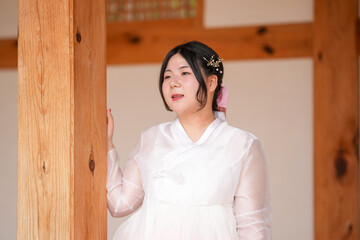 A Korean woman in her twenties wearing a charming pink hanbok peeks from the shadow of a wooden...