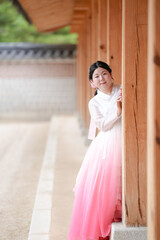 A Korean woman in her twenties wearing a charming pink hanbok peeks from the shadow of a wooden...