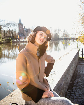 Fashionable woman in winter coat berett and reading glasses sitting in front of spectacular view of water canals going through Bruges in Belgium and  Church of Our Lady originally  Onze-Lieve-Vrouweke