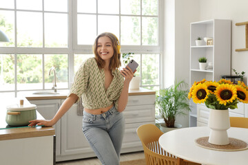Cool young woman with mobile phone listening to music and dancing in kitchen