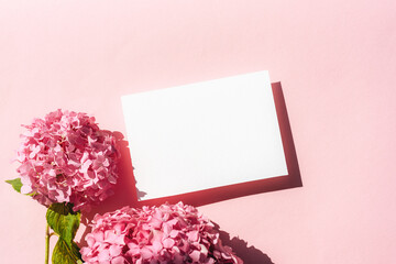 Blank white card with pink hydrangea flowers on pastel pink background with hard shadows. Top view, flat lay, mockup