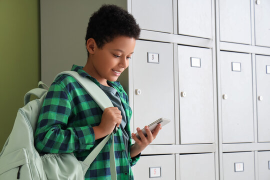 Teenage African-American schoolboy using mobile phone near locker on green background