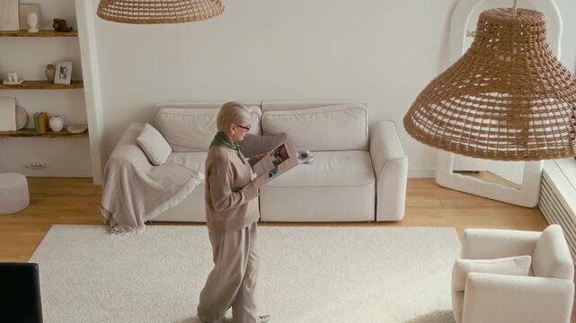 Tilt down from above shot of pensive mature Caucasian woman strolling back and forth in her living room with magazine in hand in calm, sunlit home environment