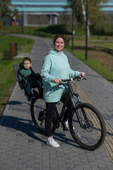 Obraz premium Caucasian woman riding bicycle with toddler son sitting behind her in child seat in park. Vertical photo. 