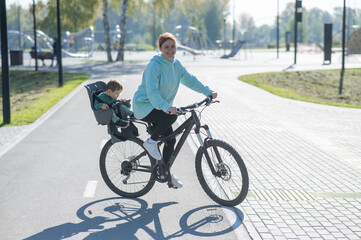 Fototapeta premium Caucasian woman riding bicycle with toddler son sitting behind her in child seat in park. 
