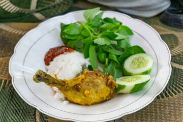 A plate contains steamed white rice, fried chicken, sliced cucumbers (Cucumis sativus), fresh green leafy vegetables, and a serving of spicy red sambal
