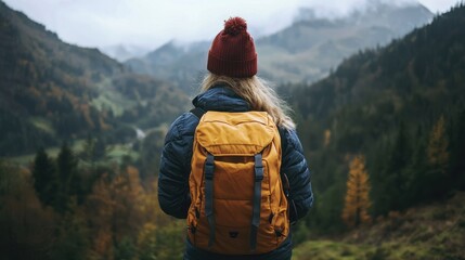 A woman in a red beanie and blue jacket with an orange backpack standing in front of a mountainous landscape with autumn colors.