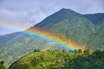 Vibrant rainbow arcs gracefully over lush green mountains, creating a stunning natural spectacle amidst serene landscape over Ulba Falls in Banos de Agua Santa, Ecuador, evoking sense of wonder