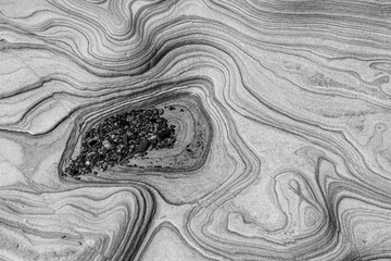 Monotone image of layered sandstone with flowing lines and a small cluster of pebbles embedded in a...