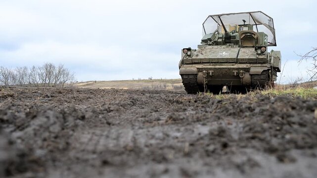 A low-angle shot of a US-made M2 Bradley on muddy terrain in Ukraine. It's adapted with a medical cross for CASEVAC and cage armor for defense against enemy drones on a muddy terrain.