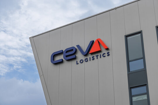 The "CEVA LOGISTICS" logo, with blue text and a striking red "A" and chevron, is mounted on the sleek, grey facade of a modern building against a bright, cloudy sky.