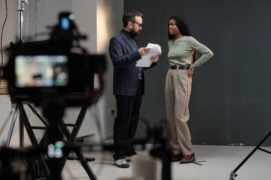 Caucasian man holding script instructing young adult Black woman during acting audition on film set with professional camera equipment visible in foreground