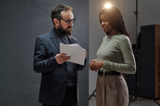 Caucasian man holding script and explaining lines to young adult Black woman during acting audition in studio setting with professional lighting equipment visible in background