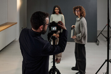 Caucasian man operating camera on tripod photographing Black young adult woman and Caucasian woman standing in studio during acting audition