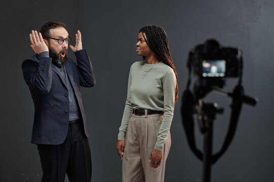 Caucasian man gesturing dramatically while standing next to young adult Black woman during acting audition in front of camera on tripod against plain background