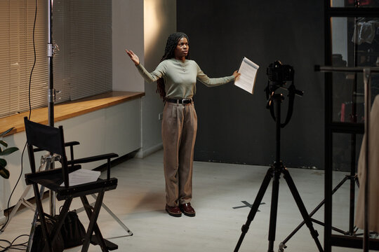 Young adult Black woman standing in front of camera holding script with arms raised, performing monologue during acting audition in studio setting, chair and tripod visible