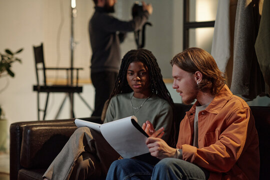 Young Black woman and young Caucasian man sitting on sofa reviewing script together, preparing for acting audition, man gesturing while discussing lines, blurred crew member in background - Powered by Adobe