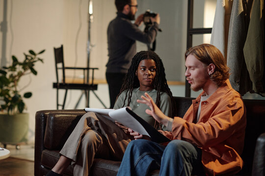 Young adult Caucasian man explaining script to young adult Black woman during acting audition while sitting on sofa, director standing in background holding camera, focused on preparation