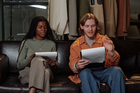 Young adult Black woman and young adult Caucasian man sitting on leather couch holding scripts, practicing lines together during acting audition, focused on reading and discussing roles