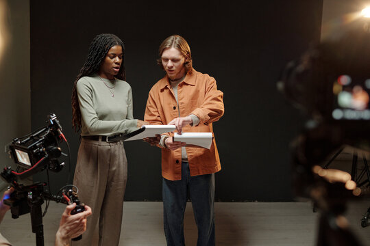 Young Black woman and young Caucasian man rehearsing script together during acting audition, standing side by side in studio while film crew recording with professional cameras