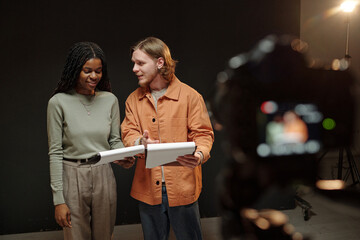 Young Black woman and young Caucasian man standing together reading scripts during acting audition, both focused on papers while camera recording in professional studio setting