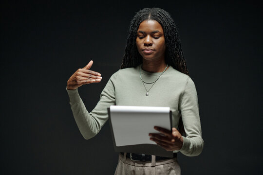 Young adult Black woman rehearsing lines during acting audition, holding script in one hand and gesturing with other hand, standing against plain dark background