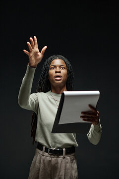 Young adult Black woman holding script, raising hand and reciting lines during acting audition, standing against dark background, expressive facial expression, focused on performance