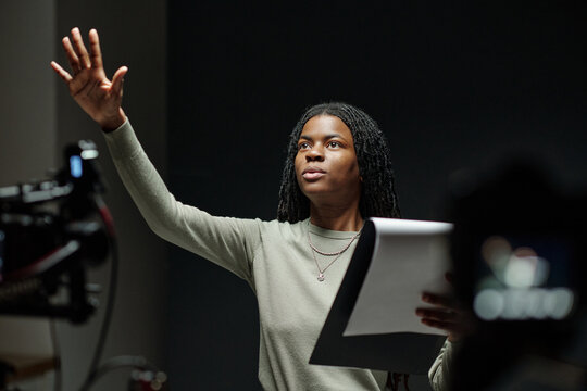Young adult Black woman holding script performing dramatic monologue during acting audition on stage, raising hand and looking upward, professional camera equipment visible in foreground