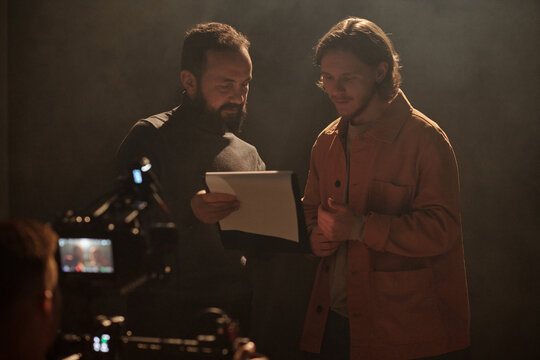 Caucasian man holding script and explaining lines to Caucasian young adult man during acting audition while professional camera operator filming scene in studio setting
