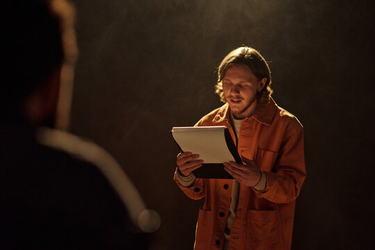 Young adult Caucasian man standing on stage reading script during acting audition, holding papers and performing lines with focused expression in front of casting director