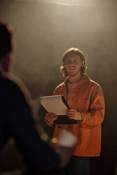 Caucasian young adult man standing on stage holding script, auditioning for acting role while looking toward casting director, facial expression showing focus and anticipation