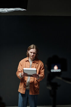 Caucasian young adult man standing in front of camera holding script reading lines during acting audition in studio setting with dark background and professional lighting equipment visible