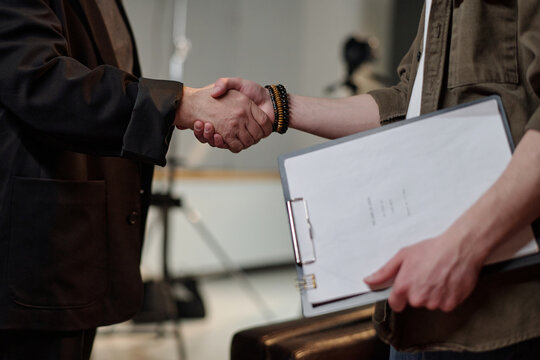 Caucasian young adult man holding clipboard with script shaking hands with middle aged Caucasian man during acting audition in studio setting, both partially visible