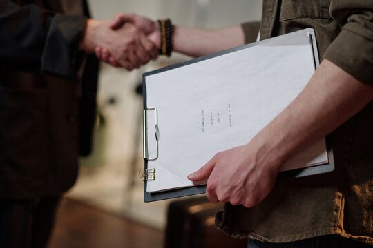 Young adult Caucasian man holding clipboard with script while standing near middle aged man shaking hands during acting audition process in professional setting