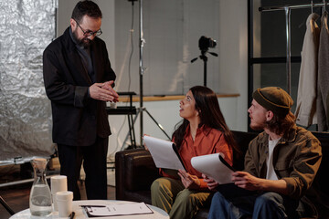 Middle aged Caucasian man instructing young adult Hispanic woman and young adult Caucasian man holding scripts during acting audition in studio setting