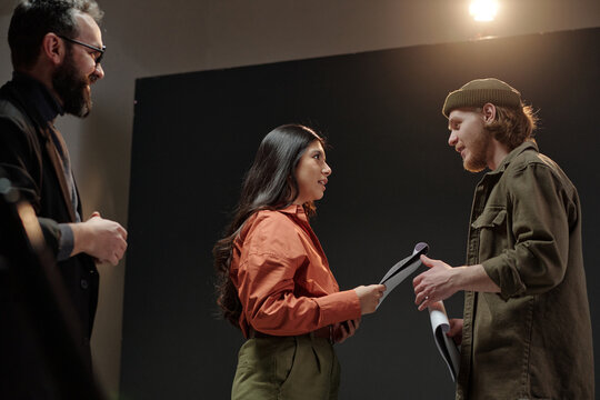 Young adult Caucasian man and young adult Hispanic woman standing face to face holding scripts engaging in acting audition while middle aged Caucasian man observing nearby