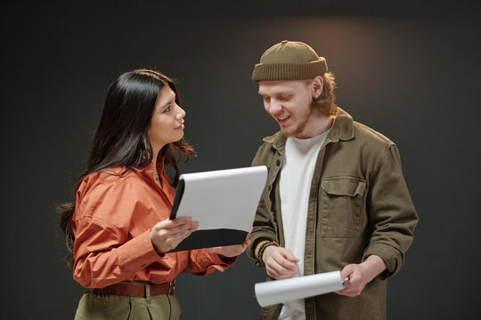 Young adult Hispanic woman and young adult Caucasian man rehearsing script together during acting audition, both holding papers and engaging in conversation against plain background