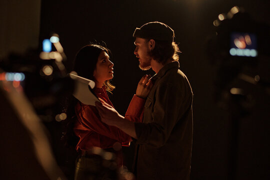 Young adult Caucasian man and Hispanic woman rehearsing dramatic scene together during acting audition, holding script and maintaining intense eye contact on dark stage