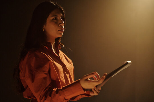 Young adult Hispanic woman rehearsing script during acting audition, holding tablet in hand, standing under stage lighting, expressing emotion while performing lines for casting