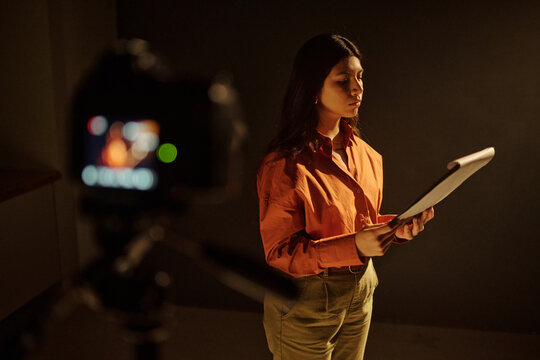 Young adult Hispanic woman standing in front of camera holding script and reading lines during acting audition, focused expression, professional studio setting, audition process
