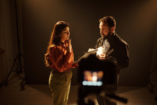 Young adult Hispanic woman rehearsing script with middle aged Caucasian man during acting audition, both standing under studio lights with camera equipment visible in foreground