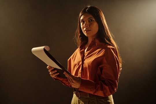 Hispanic woman holding script and rehearsing lines during acting audition, standing against dark background with focused expression, preparing for performance