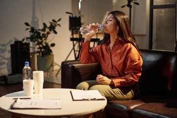 Hispanic woman sitting on sofa drinking water during acting audition preparation, holding bottle with casting documents and disposable cups on table in front