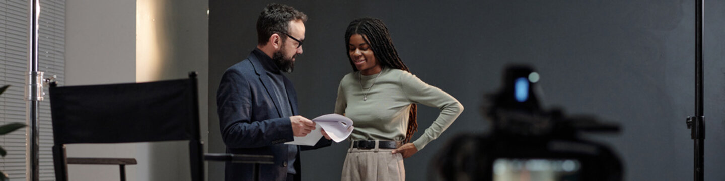 Caucasian middle aged man holding script and talking with young adult Black woman during acting audition on studio set, both standing near director chair and professional video camera