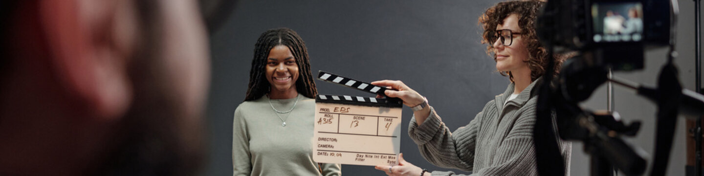 Black girl standing in front of camera smiling during acting audition while Caucasian woman holding clapperboard, professional film equipment visible in studio setting