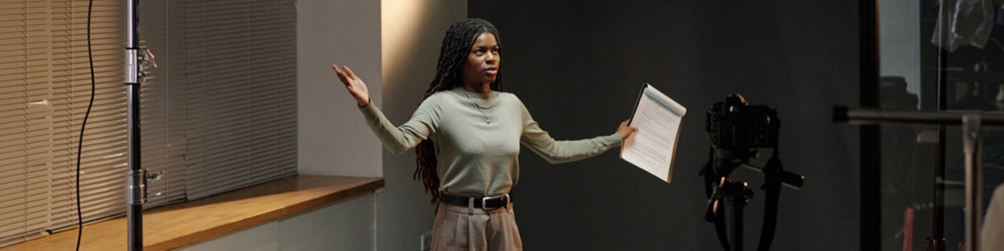 Black young adult woman standing in front of camera holding script, gesturing with arms while performing monologue during acting audition in professional studio setting