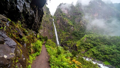 A rugged trekking trail runs parallel to a cascading waterfall tumbling down a rocky mountain